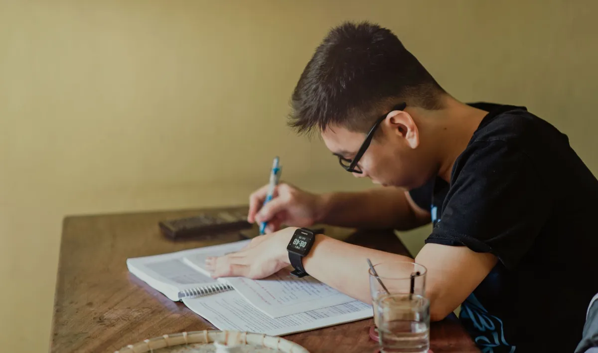A young man concentrates on studying at his desk, taking notes indoors.